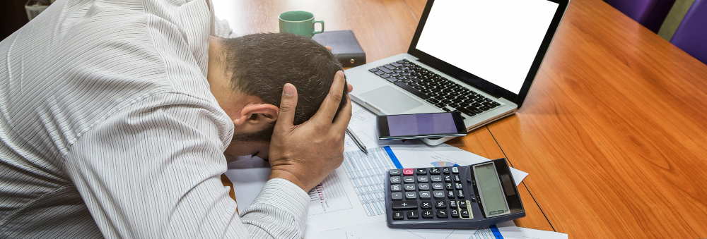 A person is seen looking worried, resting their head on the table while calculating expenditure costs, with a laptop and devices nearby.